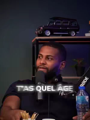 A man is sitting in front of a microphone in a podcast setting. The overlaying text reads 'T'AS QUEL ÂGE'. The background is a dark room with various objects such as a water bottle, a small replica car and a mini french fries container.
