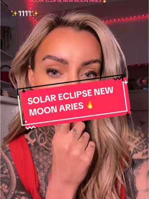 A woman with blonde hair and tattoos is the central focus. She is looking at the camera, with a red rectangular text overlay over her face. The text says “Solar Eclipse New Moon Aries” with a flame emoji. The background is softly lit, and there are strings with lights.