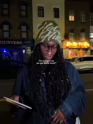 The cover image shows a person standing in front of a city street at night. They're looking down, holding a pad or device. Behind them, the street is flanked by buildings with illuminated windows and storefronts, one of which has a illuminated sign that reads "HUNTERS". The person is wearing eyeglass, a green hat, and dark clothing. Text overlay stating "'I'm hungry can we get some food?'" is added.