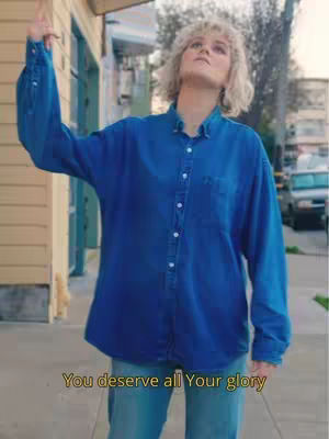 A woman with blonde hair wearing a blue denim shirt and jeans stands outside, raising her right arm upwards towards the sky. Text at the bottom of the image reads, "You deserve all Your glory."