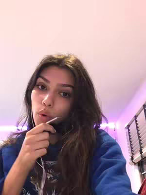 A young woman with long brown hair looks directly at the camera, holding a phone near her mouth as if ready to speak. She is wearing a blue sweater, and the background gives a glimpse of a room with a soft pink and purple lighting.