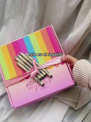A hand holding a brightly colored box with a rainbow stripe design. The box is tied with a pink ribbon, and pens are on top. A handwritten tag is also attached to the front of the box. The background is soft and neutral.