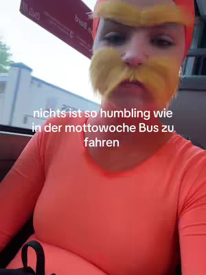 A person wearing an orange shirt with a yellow wig and mustache, taking a selfie on a bus. The background is blurred, showing the interior of the bus and the outside world.