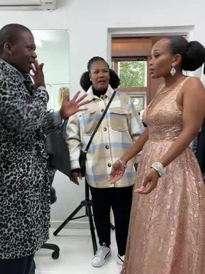 The image shows three people in what appears to be an indoor setting, likely a room. The composition is roughly a medium shot. The focus is on two women and a man. The lighting seems to be from overhead and from the right side of frame. The woman on the right side has a gold dress on. Another woman is the center next to the man on the left. In the background, there is a mirror, a doorway, and some lighting setup equipment.