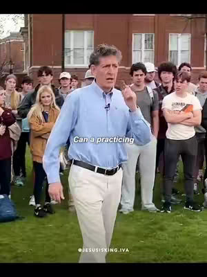 A middle-aged man in a light blue button-down shirt and off-white pants stands in front of a crowd of people, likely outdoors. The backdrop includes a brick building and some trees. The man is gesturing with his right hand and appears to be speaking. The lower portion of the image has the text overlay, "Can a practicing".