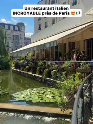 An outdoor scene shows a restaurant with a canal in the foreground. Text overlay at the top of the image indicates "Un restaurant italien INCROYABLE près de Paris". The restaurant has a white awning and looks inviting. People are seen sitting at tables under the awning. A canal with lily pads and green plants runs in front of the restaurant. The sky is blue with slight clouds.