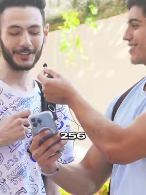 Two young men are in an outdoor setting. One of the men is holding a phone and a microphone and is speaking to the other man. There is an overlay numerical text in the image. The background is a blurry green.