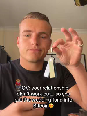 A man in a black shirt with a small emblem on the chest holds up a keychain with three colored tags. The text overlay reads, 'POV: your relationship didn't work out... so you put the wedding fund into Bitcoin'.