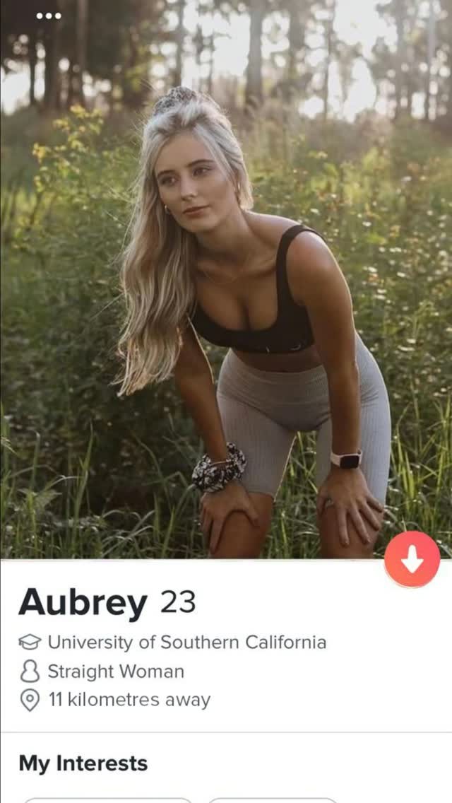 The image is a profile from a social media app. The primary subject is a fair-skinned woman with long blonde hair. She is wearing a black top and grey shorts, leaning slightly forward with her hands on her knees. The background appears to be outside, as there is a green and slightly blurred appearance of the natural environment. At the top of the image are three white dots, and below the image is a display of information, including the name "Aubrey", age "23" and a few interests.