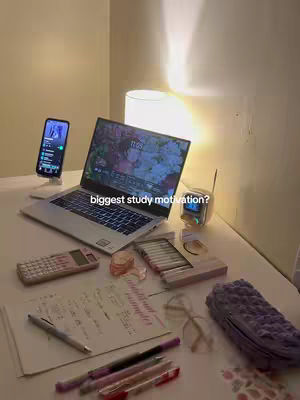 A well-lit photo shows a desk setup with study materials. A laptop with a floral screen displays, a cell phone in a stand. There are pens, papers, and a calculator next to it.