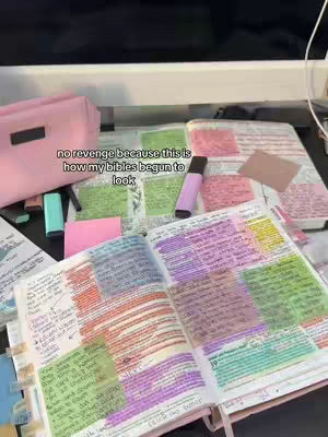 An open Bible is laid out on a desk, with pages marked up with multi-colored highlighters and sticky notes. A pink pencil case, pens and highlighters also rest on the desk.