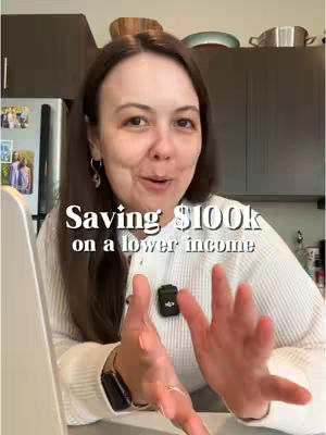 A woman is in a kitchen setting, smiling and gesturing with her hands. The overlaid text reads 'Saving $100k on a lower income'. The background includes kitchen cabinets, a refrigerator with photos, and various kitchen items.