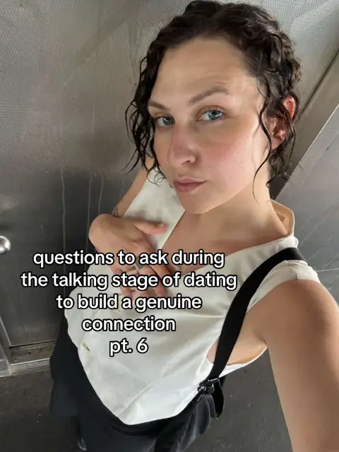 A selfie-style shot taken in what appears to be a public transit setting. The main subject is a person with curly dark hair, fair skin, and blue eyes. They are wearing a white top with a black strap. The background is a textured metallic wall. Overlayed text in white, reads "questions to ask during the talking stage of dating to build a genuine connection pt. 6".