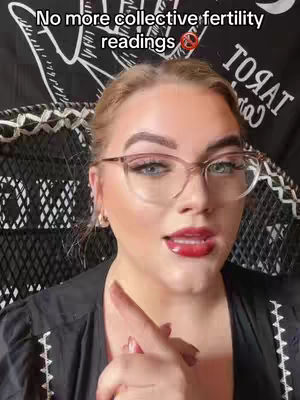 A woman with blonde hair and glasses points her index finger forward while speaking. The background includes a patterned fabric and text. The woman is wearing lipstick and a black top.