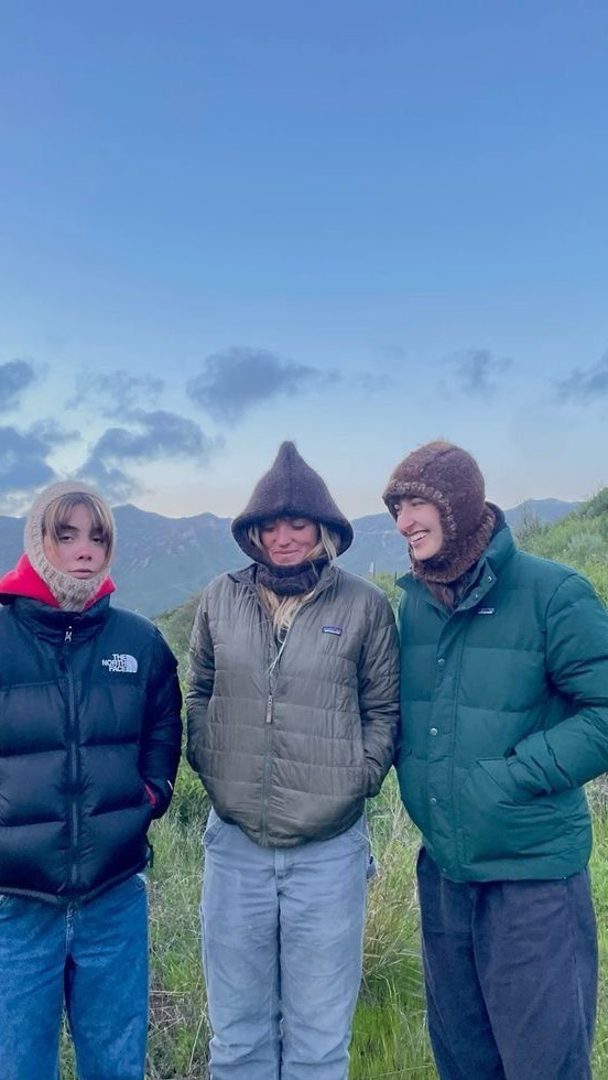 Three people are standing together outdoors, likely in a mountainous area, and facing the camera. The person on the left wears a black puffer jacket over a red hoodie and blue jeans, with a knitted hat. The person in the middle has a hooded jacket, light blue pants, and a hat. The person on the right wears a green puffer jacket and blue pants, also with a hat. The background shows a blue sky with some clouds and distant mountains.