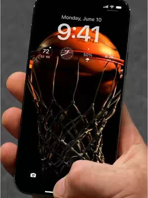 A hand holding a black iPhone displaying a basketball hoop with a basketball inside as a live wallpaper. The phone screen shows the current time, date, and battery percentage. The background is a dark gray.