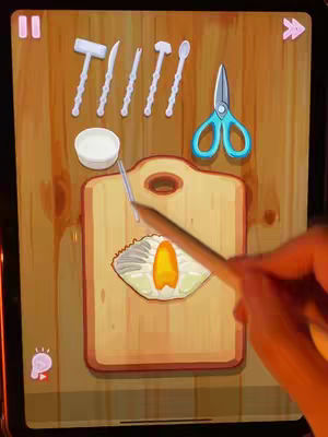 A tablet screen displaying a cooking game interface. The background appears to be a wooden table with game assets overlaid. A hand holding a stylus is interacting with the screen, and various cooking utensils and ingredients are depicted.