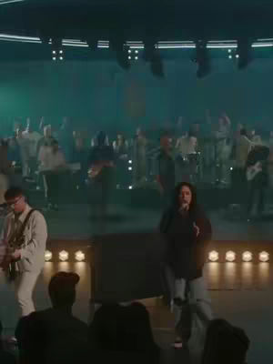 The cover image shows a church performance in a teal-lit environment. A woman with dark hair, wearing a black hoodie and light-colored pants, stands center stage, holding a microphone and singing. Other people are visible in the background, on a stage with lights. The colors are muted with a focus on teal and gold lights.