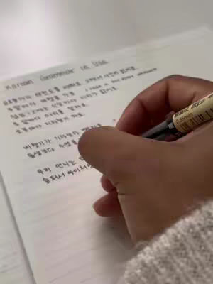 A close-up shot of a person's hand writing on a piece of lined paper with a black pen held in their hand. The paper contains handwritten Korean text and is positioned on a white surface. The person is wearing a white sweater, portion of which is visible.