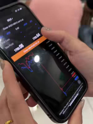 A close-up shot shows a woman's hands holding a smartphone angled towards the viewer. The phone's screen displays a trading app with a financial chart and numerical data. The woman's nails are painted, and a ring is visible. The background is out of focus, with hints of other people and furnishings.