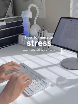The cover image shows a white desk featuring a keyboard and an iPad displaying text. White text overlays read "how to manage stress" and "over difficult work loads". There is a desk lamp and various desk supplies and a computer monitor.