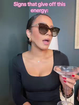 A woman is shown in the image wearing large sunglasses, a black top, and holding a cocktail glass. The background includes a glimpse of a dark wall and a light-colored lamp. Text overlay indicates the video is about astrology and zodiac signs.