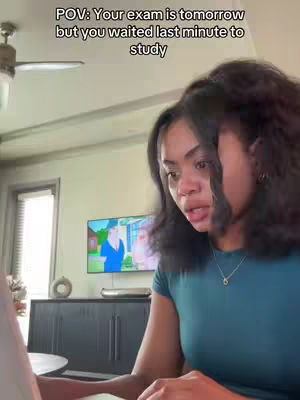 A young woman with dark curly hair sits at a table, looking at a laptop screen. A ceiling fan is visible above her, and a television and furniture are blurred in the background. Text overlaid on the image reads, 'POV: Your exam is tomorrow but you waited last minute to study'.