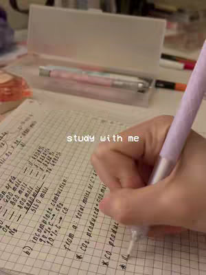A close-up shot shows a hand holding a purple pen and writing on a grid-lined paper filled with equations. In the background, a clear pencil case and other stationery items are visible, suggesting a study environment.