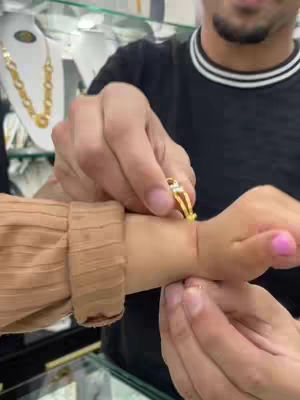 A man's hands are holding a baby’s hand. The man is placing a gold bracelet with a small diamond on the baby's wrist. The background is a jewelry store.