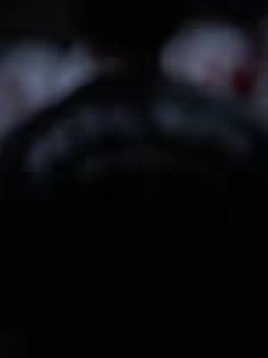 A very dark, blurred image. Shadows and indistinct shapes dominate, with a suggestion of a human figure visible, possibly a face or upper body, against a less defined and lighter background. The overall impression is one of obscurity and mystery. Colors are very subdued, leaning toward blacks, dark grays, and perhaps hints of other darker hues.