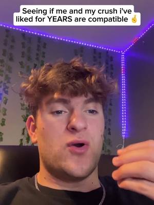 A young man with light skin and short brown hair is centered in the frame, looking directly at the camera. The background shows a wall with leafy green decorations and purple LED strip lights. The man wears a black t-shirt and a silver chain, with his hand partially in frame.