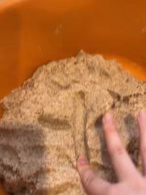 A close-up shot of a hand touching a textured brown surface, possibly bread or a similar food item. The surface fills most of the frame, with an orange background visible at the top. The lighting is warm, and the composition is focused on the tactile experience.
