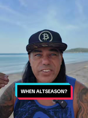 A man wearing a black baseball cap with a Bitcoin logo smiles towards the camera on a beach. A text overlay in a colorful rectangle reads: WHEN ALTSEASON?