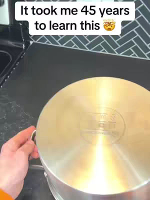 A person's hand holding the handle of a large, shiny, silver pot lid. The lid is centered in the frame, reflecting light, likely in a kitchen setting, with a dark surface below.