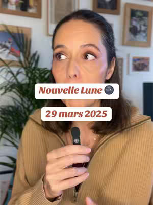 A woman with brown hair and makeup looks slightly off-camera, holding a black microphone. Text overlays read \"Nouvelle Lune\" and \"29 mars 2025\". The background includes framed pictures on a white wall, and a green plant is visible on the left.