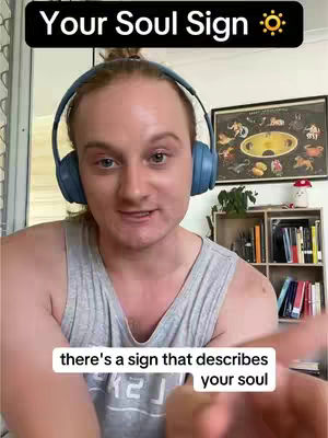 A man with light skin and reddish-blond hair, wearing a gray tank top and blue headphones, looks directly at the camera. Text overlays appear at the top, reading “Your Soul Sign” with a sun icon. A smaller text box at the bottom of the screen reads “there's a sign that describes your soul”. In the background, a bookshelf with decorative items and a framed astrology chart is visible.