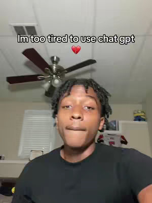 A young Black man with short dreadlocks is looking at the viewer with a tired expression. He is wearing a black shirt. In the background is a ceiling fan and the top of a bookshelf.