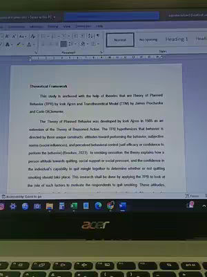 A laptop screen displaying a Microsoft Word document titled "Theoretical Framework." The document is a research paper, clearly visible with text and formatting. The laptop is on a desk with a blurred background.