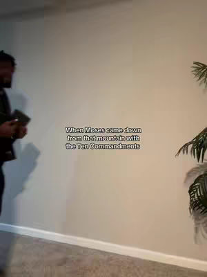 A man stands to the left holding a book, facing the right. Text overlays the image, a plant is visible to the right and part of a wall in the background