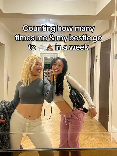 Two young women are taking a mirror selfie in a well-lit hallway, with the overlaid text. The woman on the left has blonde hair and is wearing a grey crop top and white pants, her hand over her stomach. The woman on the right has dark hair, a white crop top, a black vest, and pink pants. They are both smiling at the camera. The overall composition is simple with a casual vibe.