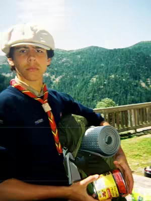 A young man wearing a scout uniform and a white newsboy cap stands in front of a mountainous, green landscape. He carries a backpack, a rolled-up sleeping pad, and a can. The scene suggests a camping or outdoor adventure. The lighting is bright and natural.