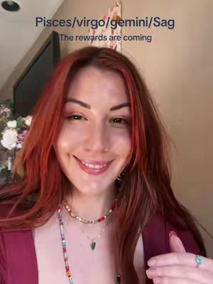 A selfie-style shot featuring a woman with red hair and a smile. The background is a blurred room interior. Text overlay lists astrological signs.