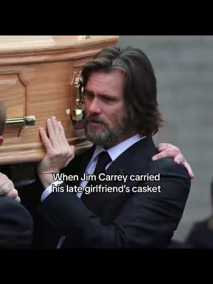 The image shows Jim Carrey carrying a wooden casket. He appears emotional, with a somber expression. The background is blurred, suggesting a funeral setting.