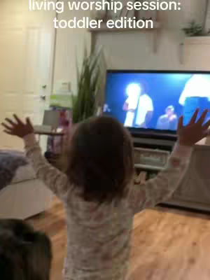 A young child, viewed from behind, stands in a living room with arms raised in a worshipful pose. The child is wearing patterned pajamas. Behind the child, a large television screen displays a performance scene. The room's background includes a door, plants, and what appears to be a bed.
