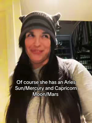A woman is wearing a gray striped hat with ear-like projections and a gray shirt. Her long black hair frames her face as she smiles. Text overlayed gives an astrology reference.