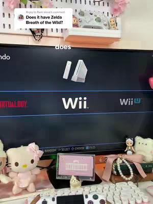 The image shows a person's desk setup with a large screen displaying the Wii interface. The desk is adorned with several plush toys, a keyboard, and a game controller. On the top right of the screen is a comment that says Does it have Zelda Breath of the Wild?.