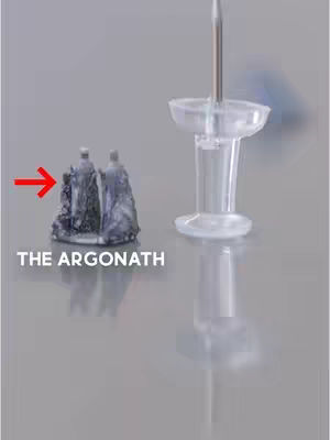 The image shows a miniature model of two figures on a gray surface, with a large push pin to the right. A red arrow points to the miniature, and white text at the bottom reads "THE ARGONATH."