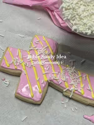 The image displays several cross-shaped cookies, frosted in pink, with yellow stripes and white sprinkles. The cookies are on a light-colored surface scattered with sprinkles. In the background, there's pink frosting in a piping bag.