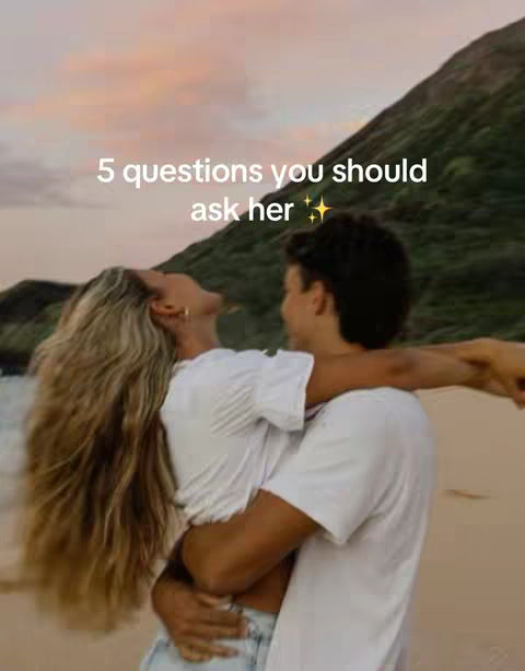 A couple embracing on a beach at sunset. The woman has long, flowing blonde hair, and the man is wearing a white t-shirt. They are set against a backdrop of a sandy beach, tranquil ocean, and mountain. The sky shows orange and purple tones.