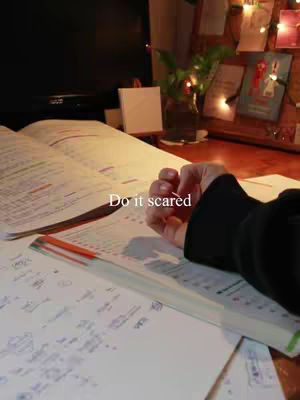A dimly lit scene with an open book on a desk. A hand is placed on top of the book, and there is a pen and some papers. The text overlay reads, 'Do it scared.'